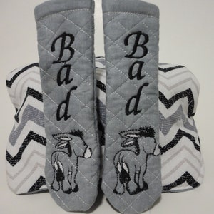 Puede incluir: Un par de guantes de horno acolchados grises con burros bordados en negro y la palabra "Bad" en cada guante. Los guantes están sobre un fondo de patrón de chevron blanco y negro.