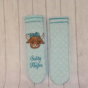 May include: A pair of teal blue quilted pot holders with white polka dots. One pot holder features an embroidered brown Highland cow with a blue bandana and the text "Salty Heifer".