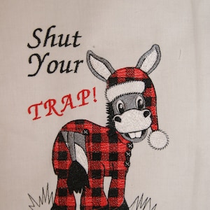 May include: Embroidered design of a donkey wearing a red and black plaid pajama top and a red Santa hat with a white pom-pom. The donkey is standing on green grass and the text "Shut Your TRAP!" is above the donkey.
