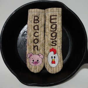 Op de afbeelding: Twee bruine, gestikte ovenwanten met geborduurde ontwerpen. Een ovenwant heeft een varken met het woord "Bacon" geborduurd. De andere ovenwant heeft een kip met het woord "Eggs" geborduurd.