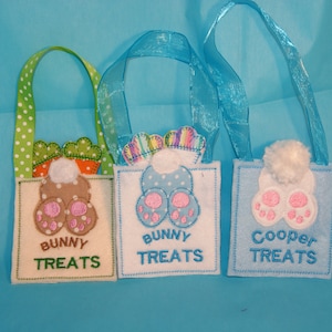 May include: Three felt bunny treat bags with bunny foot designs. The bags are light blue, white, and tan. The bags have green, white, and blue ribbon handles. The bags are labeled "Bunny Treats" and "Cooper Treats".