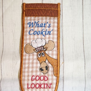 May include: A brown quilted pot holder with an embroidered moose wearing a chef's hat. The text "What's Cookin' Good Lookin'" is embroidered on the pot holder.