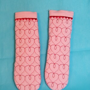 May include: A pair of pink fabric pot holders with a red heart pattern. The pot holders have a scalloped edge.
