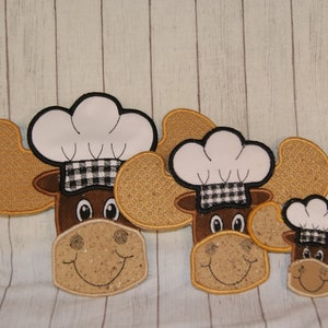 May include: Three embroidered moose heads wearing chef hats. The moose heads are brown with white chef hats and black and white checked bands. The moose heads are different sizes.