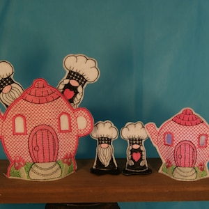 Puede incluir: Tres casas en forma de tetera de color rosa y blanco con gnomos que llevan gorros de cocinero. Las casas tienen puertas y ventanas rojas. Los gnomos están de pie frente a las casas.