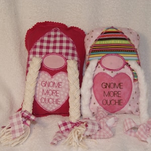Può includere: Due bambole gnomo rosa e bianche con trecce bianche. Gli gnomi indossano cappelli a quadri rosa e bianchi e a righe. Gli gnomi hanno cuori rosa sul petto con il testo "Gnome More Ouchie".