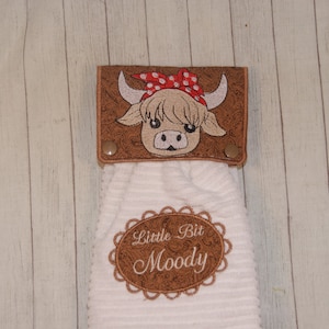 May include: White kitchen towel with a brown embroidered cow head with a red polka dot bandana and the text "Little Bit Moody" in a brown oval.
