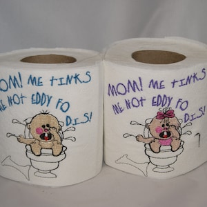 Puede incluir: Dos rollos de papel higiénico con diseños bordados. Un rollo presenta un bebé con el texto "MOM! ME FINKS ME NOT EDDY FO DIS!" El otro rollo presenta una niña con el texto "MOM! ME TINKS ME NOT EDDY FO DIS!"