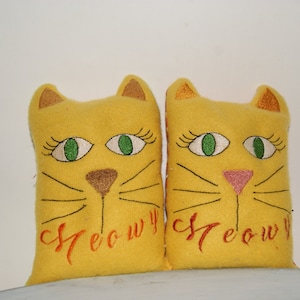 May include: Two yellow felt cat pillows with embroidered green eyes, brown noses, and black whiskers. The pillows have the word "Meow" embroidered on them in orange thread.