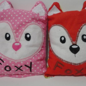 May include: Two plush fox pillows, one pink with white polka dots and the other red, both with embroidered white faces and the word "Foxy" on the bottom.