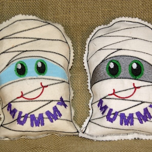 May include: Two white fabric mummy pillows with embroidered eyes, mouths, and the word "MUMMY" on each. One mummy has blue eyes and the other has gray eyes.