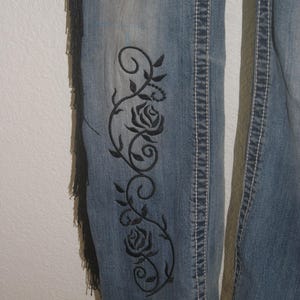 May include: Light blue denim jeans with black embroidered roses and vines on the side. The jeans have white stitching and a fringed detail on the side. The jeans are a classic style.