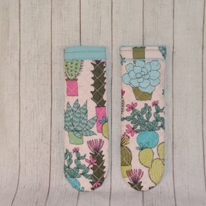 May include: Two fabric oven mitts with a light pink background and a cactus pattern. The mitts feature various cacti in pots, with green, pink, and blue accents. The top edges are trimmed with a light blue fabric.