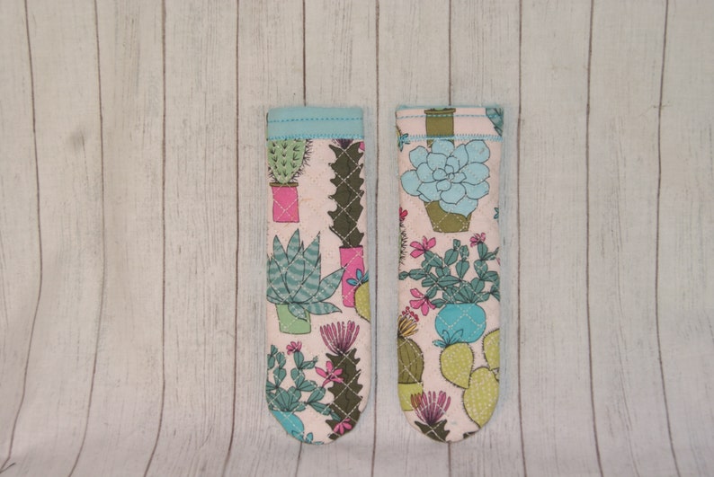 May include: Two fabric oven mitts with a light pink background and a cactus pattern. The mitts feature various cacti in pots, with green, pink, and blue accents. The top edges are trimmed with a light blue fabric.