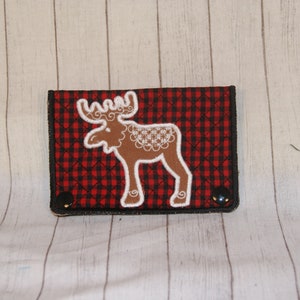 May include: A red and black plaid wallet with a brown moose applique. The moose has white decorative accents. The wallet is rectangular with black trim and two black snap closures.