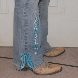 Puede incluir: Vaqueros de mezclilla azul claro con plumas turquesas bordadas y el texto "COWGIRL UP" en blanco. Los vaqueros se combinan con botas de vaquero color canela.