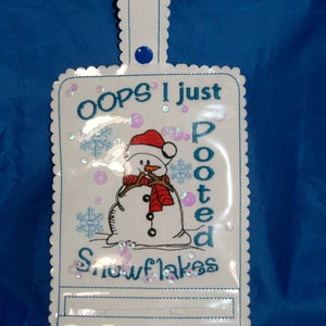 Pode incluir: Ornamento de tecido branco com um desenho de boneco de neve e o texto "OOPS I just pooted snowflakes". O ornamento tem um botão azul e um laço para pendurar.