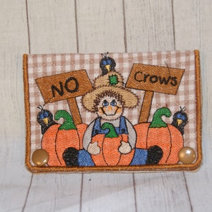 May include: A brown and tan fabric wallet with an embroidered image of a scarecrow holding pumpkins and a sign that says "No Crows".