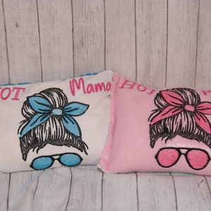 May include: Two embroidered pillows with the text "Hot Mama" and "Hot Mama" on them. The pillows are white and pink and feature a cartoon image of a woman with a messy bun and sunglasses.
