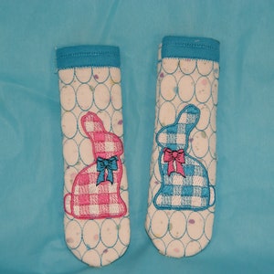 Könnte beinhalten: Zwei Topflappen aus Stoff mit weißem Hintergrund und einem blauen und weißen Gingham-Hasendesign. Ein Hase ist rosa und der andere ist blau.