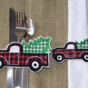 May include: Two Christmas-themed napkin rings shaped like red and black plaid trucks carrying green and white checkered Christmas trees. One ring is around a white napkin, the other is next to silverware.