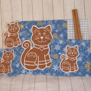 以下が含まれることがあります： 猫をテーマにしたキッチン用テキスタイルセット。セットには、プレースマット、コースター2つ、カトラリーホルダーが含まれます。各アイテムは、青い雪の結晶模様の背景に白い刺繍が施された茶色の猫のデザインが特徴です。シナモンスティックが含まれています。