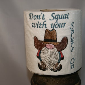 May include: A white toilet paper roll with an embroidered gnome wearing a brown cowboy hat and boots. The text "Don't Squat with your Spurs On" is embroidered on the roll.