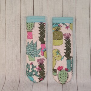 May include: Two fabric oven mitts with a light pink background and a pattern of various cacti in pots. The mitts have a light blue trim. The cacti are green, pink, and teal.