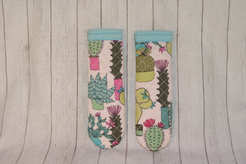 May include: Two fabric oven mitts with a light pink background and a pattern of various cacti in pots. The mitts have a light blue trim. The cacti are green, pink, and teal.