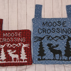 May include: Two quilted pot holders with a moose crossing design. One is red and black plaid with white embroidery. The other is blue with black embroidery and white stars. Both have a loop for hanging.