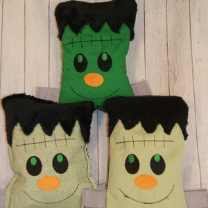 May include: Three green and black plush Frankenstein-themed pillows with embroidered faces. The pillows are arranged in a triangle shape.