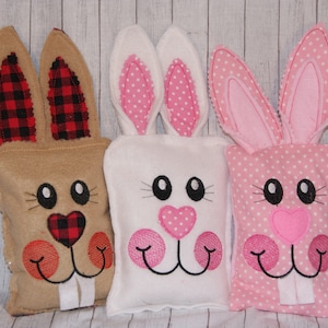 May include: Three plush bunny-shaped pillows with embroidered faces. The bunny on the left has brown fur with red and black plaid ears. The middle bunny is white with pink polka dot ears. The bunny on the right is pink with white polka dot ears.