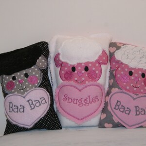 May include: Three plush lamb-shaped pillows with embroidered faces and hearts. The pillows are black and white polka dot, white with pink polka dots, and gray with pink hearts. The hearts have the words "Baa Baa", "Snuggles", and "Baa Baa" embroidered on them.