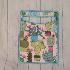 May include: A rectangular fabric card holder with a light pink background and a pattern of various cacti in pots. The holder has a turquoise border and green button closures. The cacti are in shades of green, pink, and blue.