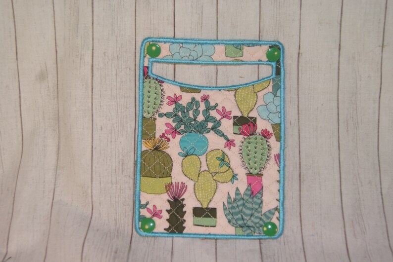 May include: A rectangular fabric card holder with a light pink background and a pattern of various cacti in pots. The holder has a turquoise border and green button closures. The cacti are in shades of green, pink, and blue.