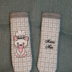 May include: Two white and blue gingham fabric pot holders with embroidered designs. One pot holder features a white cat with a pink nose and a chef's hat. The other pot holder has a white heart with the text "Meow Mix" embroidered inside.