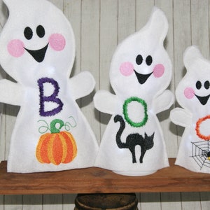 Puede incluir: Tres adornos de fantasmas de fieltro blanco con caras y detalles bordados. El primer fantasma tiene una "B" morada y una calabaza naranja, el segundo tiene una "O" verde y un gato negro, y el tercero tiene una "O" naranja y una telaraña con una araña.