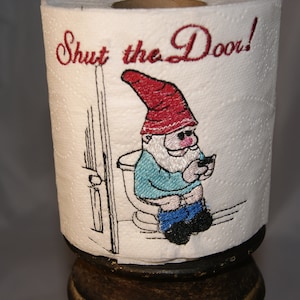 Puede incluir: Un rollo de papel higiénico blanco con un diseño bordado en rojo y negro de un gnomo sentado en un inodoro con un teléfono en la mano. El texto "Shut the Door!" está bordado en rojo sobre el gnomo.