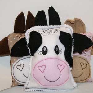 May include: Three plush cow-themed cushions with brown, white and pink fabric. The centre cushion has a black and white cow face with pink nose and eyelashes. The other two cushions have brown ears and pink polka dots.