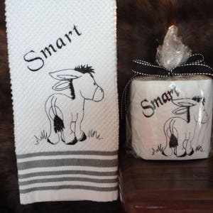 May include: A white hand towel with black embroidery of a donkey and the word "Smart". A white toilet paper roll with black embroidery of a donkey and the word "Smart" is wrapped in clear plastic and tied with a black ribbon.