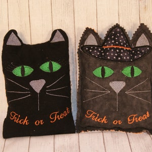 May include: Two black cat-shaped plush pillows with green eyes and gray noses. Both pillows have the text "Trick or Treat" embroidered on them in orange thread. One pillow has a black witch hat with white stars on top.