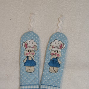 May include: Two blue and white pot holders with a quilted design. Each pot holder features an embroidered bunny wearing a chef's hat.