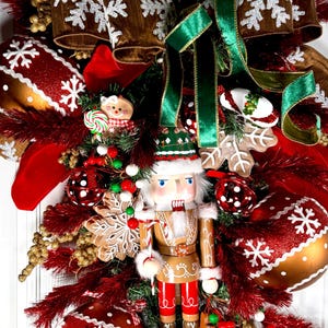 May include: A Christmas wreath with a gingerbread nutcracker, red and gold ornaments, and a brown ribbon with white snowflakes. The wreath is decorated with red and green berries, and gold glitter.