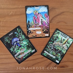 May include: Three rectangular game cards with hand-painted illustrations. One card depicts a pink insect with the text Insect, Token Creature - Insect, Flying, and 2/1. Another shows a blue fungus with Fungus, Token Creature - Fungus, and 1/1. The third shows mushrooms with Saproling, Token Creature - Saproling, and 1/1.