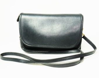 May include: A black leather crossbody bag with a flap closure and a long, thin strap. The bag has a rectangular shape with rounded edges and gold-coloured hardware. The strap is designed to be worn over the shoulder.