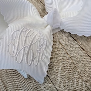 May include: A large, white grosgrain ribbon bow with a scalloped edge. The bow features an embroidered monogram in white thread. The bow is set against a wooden background. The text "Lady Bowtique" is visible in the lower right corner.