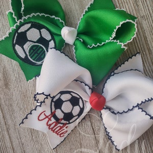 May include: Two green and white hair bows with a soccer ball design and white trim. One bow has the name "Addie" embroidered on it.
