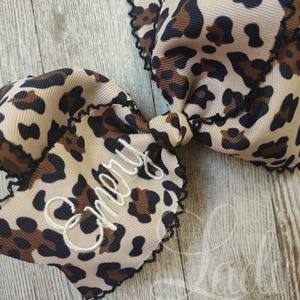 May include: A brown and beige leopard print hair bow with black trim and the name "Cheryl" embroidered in white.