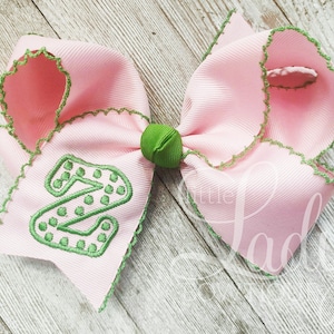 May include: A pink hair bow with a green embroidered letter "Z" with white polka dots. The bow has a green centre and a scalloped edge.