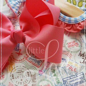 May include: A pink and white fabric with a blue and white gingham trim. The fabric has a pattern of stamps with words like "Adventure Begins", "Air Mail", and "Serendipity". A large pink bow is on top of the fabric.